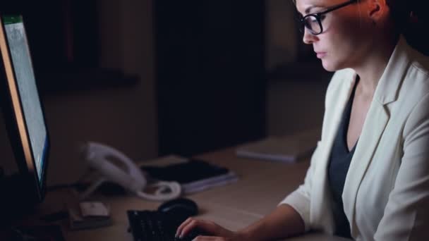 Chica de confianza en la ropa formal oficinista está utilizando la computadora tarde en la noche trabajando solo en la oficina oscura sentado en el escritorio y mirando a la pantalla. Concepto de trabajo y millennials . — Vídeos de Stock