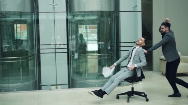 Movimento lento de caras felizes em ternos se divertindo no lobby do escritório montando cadeira rolante e jogando papéis gritando e rindo. Conceito de espaço de trabalho e alegria . — Vídeo de Stock