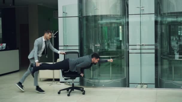 Joyful trabajadores de oficina se están divirtiendo con la silla rodante en el vestíbulo del centro de negocios sillón de montar y riendo disfrutando de la actividad divertida en la fiesta de trabajo . — Vídeos de Stock