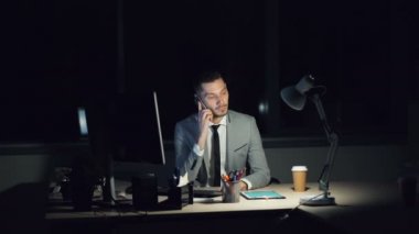Cep telefonu masada oturma gülüyor konuşurken gece geç ofisinde çalışan genç adam vurguladı. Tahrik bilgisayar, gitmek kahve görünür.