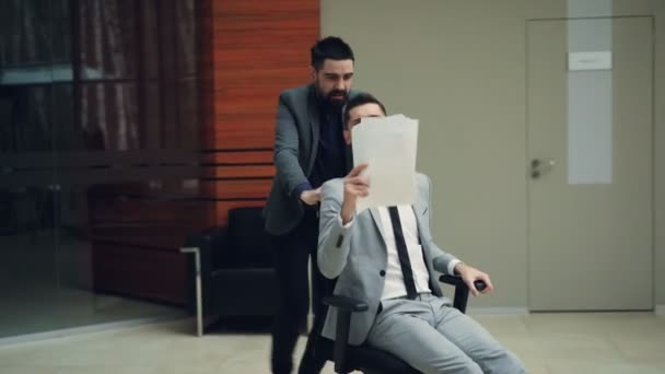 Joyeux employés de bureau masculins ont plaisir à monter chaise dans le hall et jeter des papiers parler et crier. Jeunesse heureuse, culture d'entreprise et concept de lieu de travail .