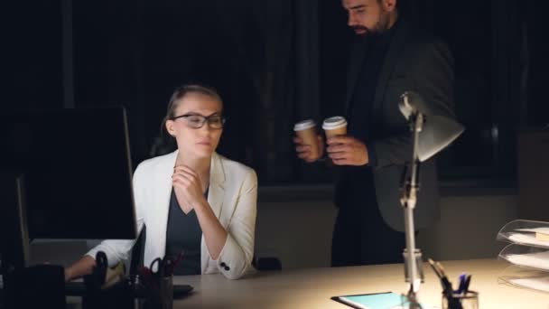 Die müde junge Büroangestellte arbeitet spät in der Nacht am Computer, als ihr Kollege ihr den To-Go-Kaffee bringt. Die Menschen unterhalten sich freundlich. — Stockvideo