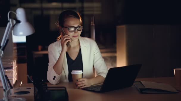 Привлекательная предпринимательница разговаривает по мобильному телефону и использует ноутбук, работающий в офисе поздно ночью. Современные технологии, трудолюбивая молодежь и коммуникационная концепция . — стоковое видео