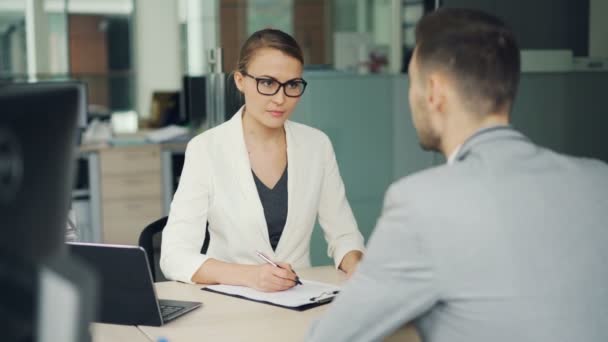 La donna d'affari amichevole in occhiali e vestito sta intervistando un candidato maschio per il lavoro in ufficio. La gente parla facendo e rispondendo alle domande, ragazza sta scrivendo . — Video Stock