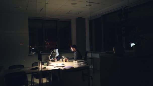 Zmęczony biznesmen w garnitur i krawat jest za pomocą komputera w biurze pracy w nocy, siedząc przy worktable w ciemnym pokoju, patrząc na ekran i pisania, a następnie picia kawy. — Wideo stockowe