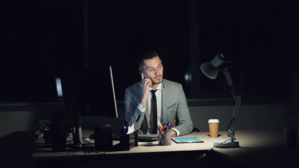Benadrukt dat de jonge man in het werken met office's avonds laat praten op mobiele telefoon dan lachen vergadering om Bureau. Gedraaid computer, en-klare koffie zijn zichtbaar. — Stockvideo