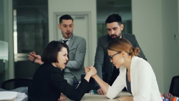 Chicas lindas trabajadores de oficina están compitiendo en la lucha libre brazo sentado en la habitación en el escritorio, mientras que los hombres jóvenes en trajes están animando a continuación, haciendo chocar los cinco. Diversión y concepto de trabajo . — Vídeo de stock