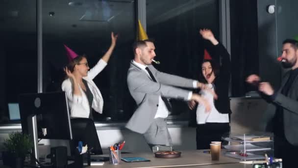 Щасливі молоді працівники офісу танцюють на вечірці в капелюхах і дме роги. Торт на день народження та подарункова коробка видно на столі . — стокове відео