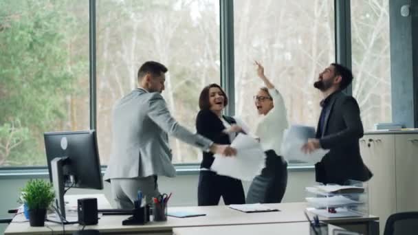 Wesoły mężczyźni i kobiety są o partii w biurze taniec z dokumentami a następnie rzucając je w powietrze i śmiejąc się. Młodzi ludzie noszą formalne garnitury. — Wideo stockowe
