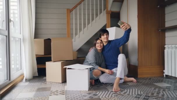 Adorable young people girl and guy are taking selfie expressing love and tenderness kissing and hugging sitting on floor of new house and using smartphone. — Stock Video