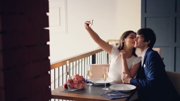 Attraktiva älskande par tar selfie med smartphone med middag i restaurangen. Unga människor ler, kyssas och poserar tittar på kameran. — Stockvideo