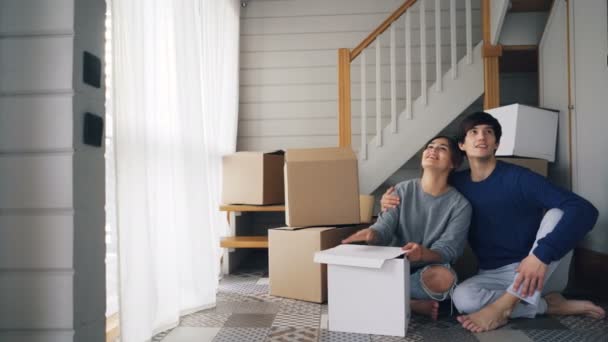 Молодые люди муж и жена разговаривают и целуются сидя на полу в новом доме рядом с лестницей и мечтают о том, чтобы строить планы на жизнь в новом доме. Переезд и молодежная концепция . — стоковое видео