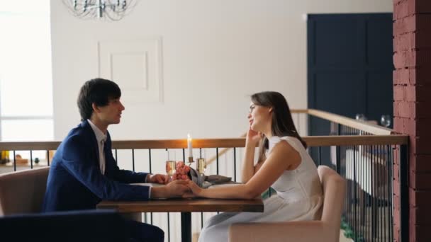 Muchacha y chico jóvenes felices están hablando tomados de la mano sentados en la mesa con flores, velas y champán en el bonito restaurante. Concepto de citas y conversación . — Vídeos de Stock