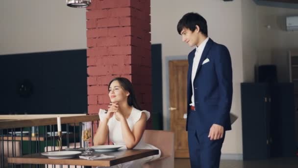 Una joven sonriente está esperando a su novio en el restaurante y luego recibe flores, lo besa y habla mientras él está sentado en la mesa frente a ella. Concepto de citas y rosas . — Vídeos de Stock