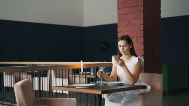 Linda joven está esperando a su novio en el restaurante sentado en la mesa, utilizando el teléfono inteligente comprobar el tiempo y luego beber vino espumoso y salir. Citas y concepto de lucha . — Vídeos de Stock