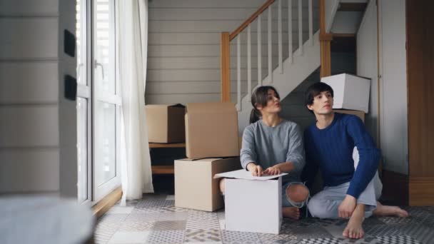 Aantrekkelijke jonge vrouw liefdevolle vriendin is in gesprek met haar vriendje dan geven hem huissleutels zittend op de vloer van nieuw appartement. Verhuizing en verhuizen in concept. — Stockvideo