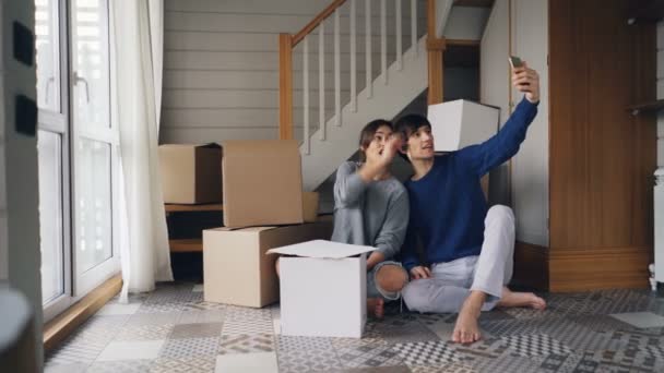 Happy couple man and woman are making online video call with smartphone after relocation. They are greeting friends, showing new house keys and boxes, chatting and smiling. — Stock Video