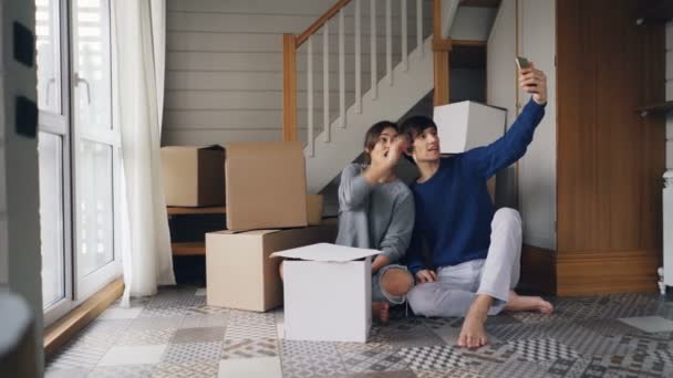 Pareja feliz hombre y mujer están haciendo videollamada en línea con el teléfono inteligente después de la reubicación. Están saludando a amigos, mostrando nuevas llaves y cajas de la casa, charlando y sonriendo . — Vídeos de Stock