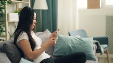 Çekici Asya Bayan smartphone evde kanepede oturan ve internette alışveriş banka kartı ile online ödeme yapmak için kullanıyor. Bankacılık ve gençlik kavramı.