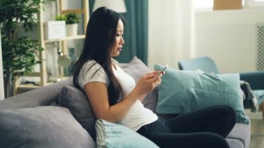 Şirin Asyalı kız öğrenci ekran dokunaklı ve evde koltukta tek başına oturmak gülümseyen modern smartphone kullanıyor. Gençlik, boş zaman zaman ve Gadget'ları kavramı.