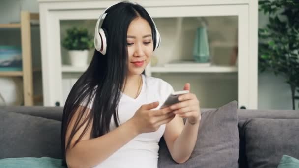 Cute young Asian woman is listening to music in headphones and touching smartphone screen resting on sofa at home. Technology, youth culture and gadgets concept. — Stock Video