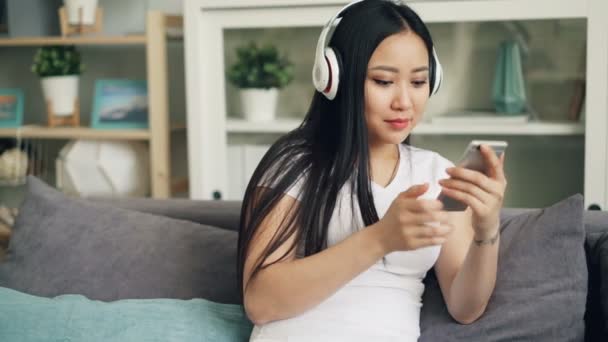 Jeune femme asiatique avec de beaux cheveux noirs longs écoute de la musique à travers des écouteurs et en utilisant smartphone profiter du temps libre à la maison. Concept jeunesse et technologie . — Video