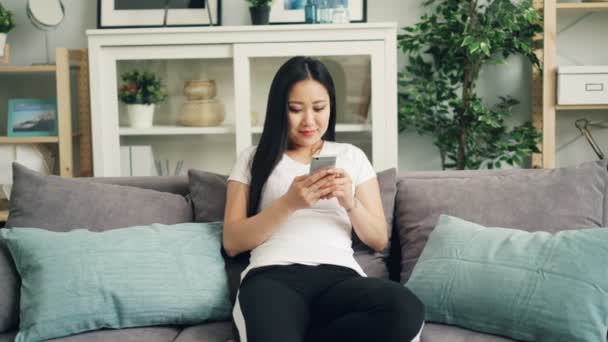 Menina asiática bonito está usando smartphone tocando tela e sorrindo relaxante no sofá em casa sozinho desfrutando de tecnologia moderna. Juventude e conceito de gadgets . — Vídeo de Stock