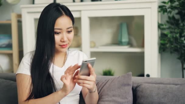 Attraktive asiatische Frau im weißen T-Shirt nutzt Smartphone Touchscreen lächelnd und lachend checkt soziale Medien und sendet SMS. Informations- und Kommunikationskonzept. — Stockvideo