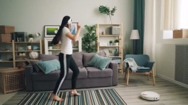 Şirin Asyalı kız öğrenci kulaklık ve robot elektrikli süpürge halı ve zemin evde temizlerken dans müzik dinliyor. Teknolojileri ve gençlik kavramı.