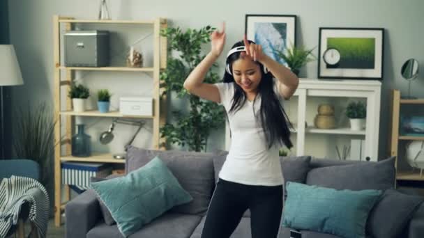 Beautiful young lady is dancing at home and singing wearing headphones and listening to music alone in living room. Modern technology, apartment and song concept. — Stock Video