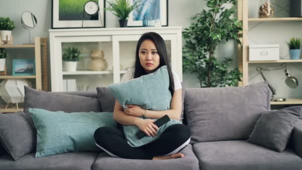 Hermosa mujer joven asiática está viendo películas de terror en la televisión en casa sosteniendo la almohada y reaccionando a los episodios de miedo a continuación, utilizando el control remoto. Concepto de películas y personas . — Vídeo de stock