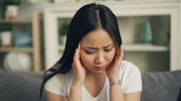 Приваблива жінка азіатських страждає від сильний головний біль, торкаючись її голова масажуючи храми в приміщенні сидячи вдома. Біль, людей і здоров'я концепції. — стокове відео