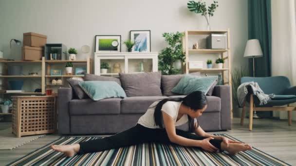 Mooie jonge dame doet fysieke oefeningen aan huis uitrekkende benen en lichaam zittend op de vloer van stijlvolle moderne appartement. Sport en millennials concept. — Stockvideo