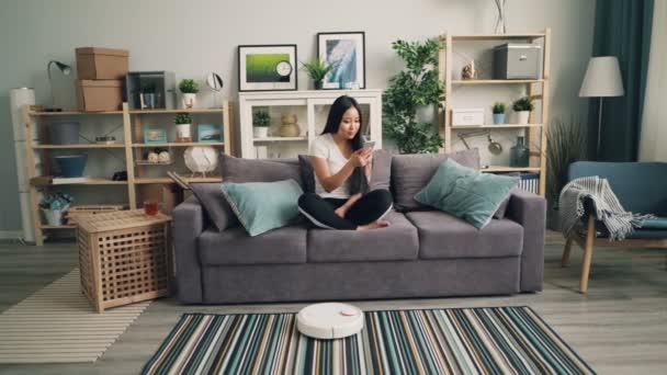Νεαρή γυναίκα της Ασίας είναι χρησιμοποιώντας το smartphone και να ακουμπά στον καναπέ, ενώ η ρομποτική σκούπα με ηλεκτρική σκούπα διαμέρισμα αντί της. Σύγχρονες τεχνολογίες και την έννοια της οικιακής. — Αρχείο Βίντεο