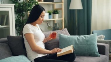 Güzel Asyalı kadın ev çay içme ve rahatlatıcı kanepede oturan modern edebiyat zevk kitap okuyor. Eğitim, millennials ve ev kavramı.