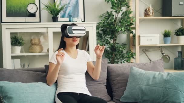 Leende ung dam i augmented reality glasögon flyttar händerna och vrida huvudet sitter på soffan hemma och ha roligt. Underhållning och teknik koncept. — Stockvideo