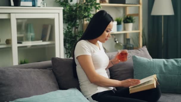 Asiatisk ung dam läser bok och innehav kopp te och sedan vänder sida och dricka sittande på soffan i fin lägenhet. Kultur och drycker koncept. — Stockvideo