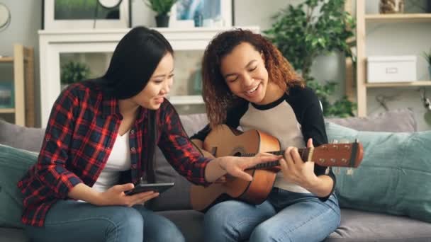 Mujer asiática está enseñando a su amigo afroamericano a tocar la guitarra durante el tiempo libre en casa. Una chica sostiene la guitarra, otra usa una tableta . — Vídeo de stock