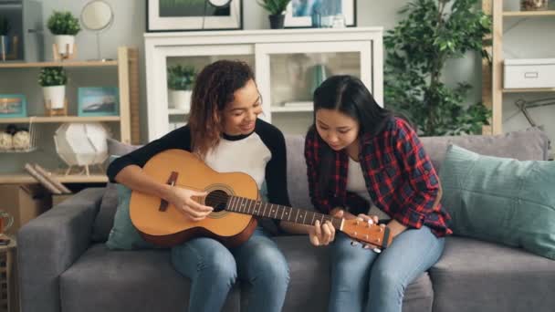 Glada afroamerikansk flicka är undervisning hennes asiatiska vän att spela gitarr hemma. Unga kvinnor sitter på soffan håller musikinstrument och talar. — Stockvideo