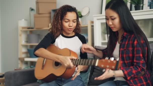 Lächelnd bringt eine Afroamerikanerin ihrem asiatischen Freund bei, die Akustikgitarre zu spielen und dann ihr Musikinstrument zu geben. Frauen reden und spielen. — Stockvideo