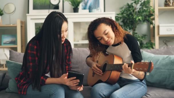 Amigos de la raza mixta asiático y afroamericano están aprendiendo a tocar la guitarra con la tableta. Las niñas están mirando la pantalla y tratando de las posiciones de los dedos en el instrumento musical . — Vídeos de Stock