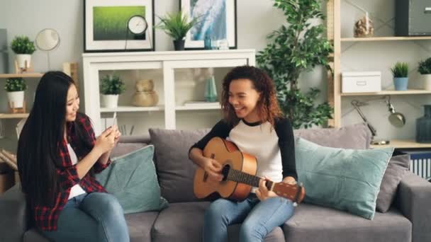 Las amigas se están divirtiendo en casa, la chica afroamericana está tocando la guitarra y la joven asiática está grabando video con teléfono inteligente. Concepto de amistad y música . — Vídeo de stock