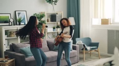 Neşeli genç kadınlarda Afrika Amerika ve Asya casual giyim gitar çalmak, dans ve Tv uzaktan kumanda modern apartman dairesinde eğleniyor şarkı.