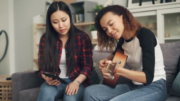 Asiatische Dame benutzt Laptop, um ihrem afrikanisch-amerikanischen Freund beizubringen, zu Hause gemeinsam Gitarre zu spielen. Mädchen reden und lachen und genießen die Aktivität. — Stockvideo