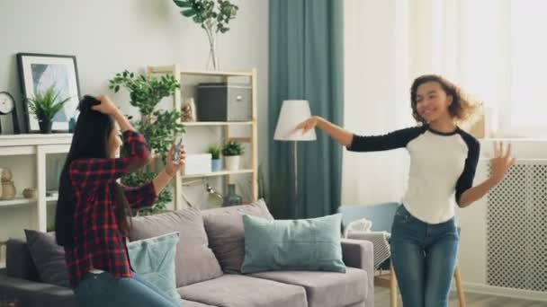 Emocional chica afroamericana estudiante está bailando en casa riendo y divirtiéndose mientras su amigo asiático está grabando vídeo con cámara de teléfono inteligente. Concepto de juventud y tecnología . — Vídeos de Stock