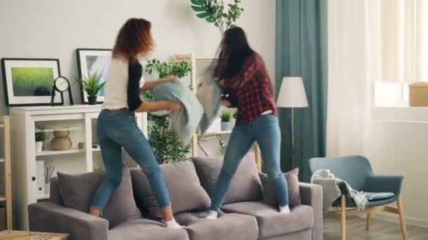 Mujeres jóvenes y juguetonas afroamericanas y asiáticas están luchando con almohadas paradas en el sofá y riendo. Las niñas están usando ropa casual camisas y pantalones vaqueros . — Vídeos de Stock