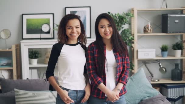 Retrato en cámara lenta de hermosas damas jóvenes asiáticas y afroamericanas de pie en casa, mirando a la cámara y sonriendo. La gente lleva ropa casual. . — Vídeos de Stock