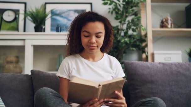 La atractiva joven afroamericana está leyendo un libro disfrutando de la literatura moderna sentada en un sofá en casa sola. Hobby, concepto de juventud e interior . — Vídeos de Stock