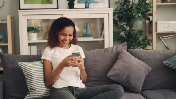 Menina afro-americana bonita está usando smartphone e rindo relaxante em sofá confortável em casa. Conceito de tecnologia moderna e estilo de vida juvenil . — Vídeo de Stock