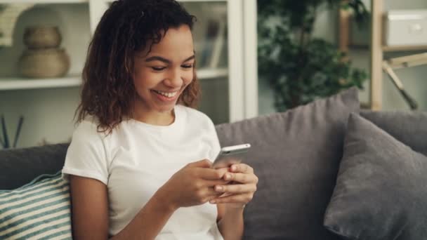 うれしそうなアフリカ系アメリカ人女性がスマート フォンを持って、画面に触れると明るい部屋でソファの上に座って笑っています。若い人、ガジェットや楽しいコンセプト. — ストック動画